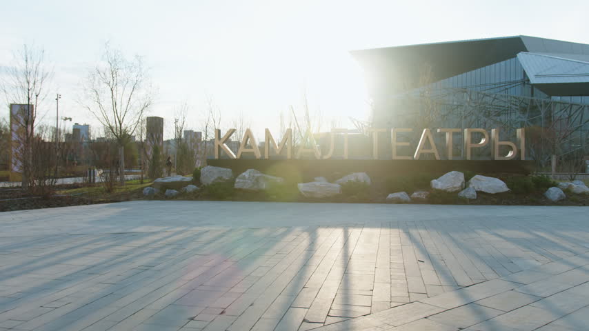 Gold theater name sign stands on paved square before modern complex. Bright sunlight shines over entrance area during calm daytime
