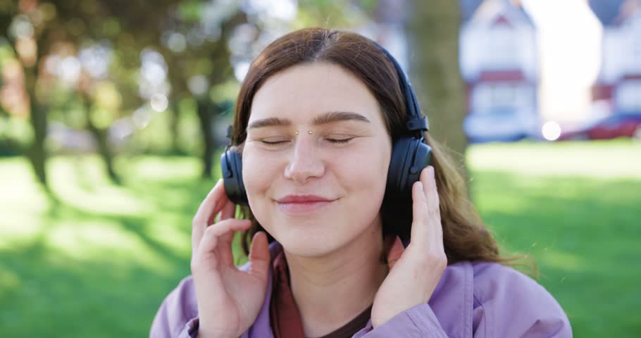 Beautiful young woman wearing headphones, sitting on a park bench, listening to music, and laughing on a sunny day in the city