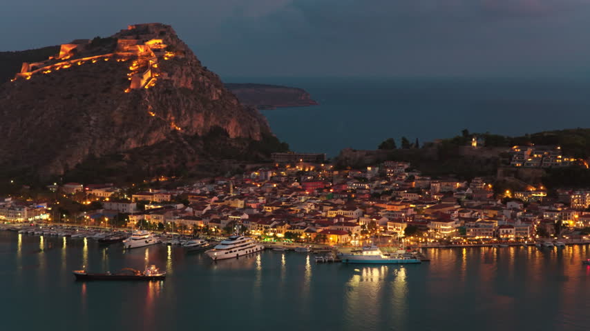 Stunning aerial view of the historic town of Nafplio in Greece, showing the illuminated Palamidi fortress and the sparkling city lights reflecting on the tranquil waters of the bay at dusk
