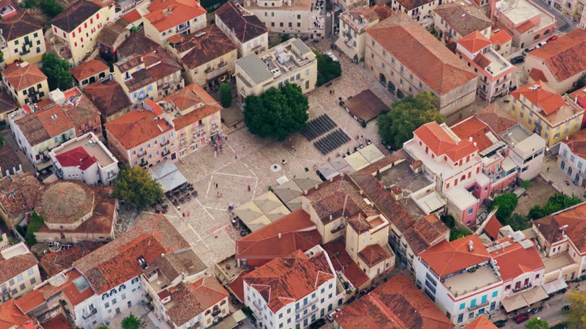 Aerial drone footage flying over the bustling Syntagma Square in the heart of Nafplio, a picturesque historic town in Greece, showcasing the traditional architecture and red-tiled rooftops