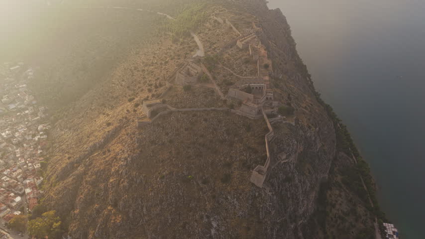 Stunning aerial perspective slowly revealing the historic Palamidi fortress and its extensive fortifications perched on a rocky hill above the city of Nafplio in Greece during a hazy sunrise