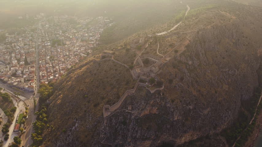 High-altitude aerial perspective showing the historic Palamidi fortress on a rocky hill overlooking the city of Nafplio in Greece, with the sun casting a warm glow over the landscape