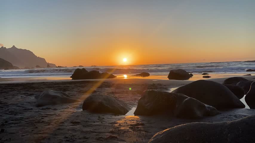 Golden sunset over the ocean with a sun flare, waves and dark volcanic boulders on a black sand beach, Tenerife, Spain