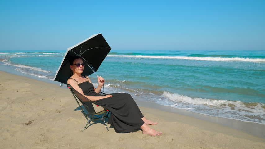 Woman relaxing on the beach with an umbrella. Solitary woman in a black dress sitting on a folding chair by the sea, holding an umbrella to shade herself from the sun while enjoying the coastal view