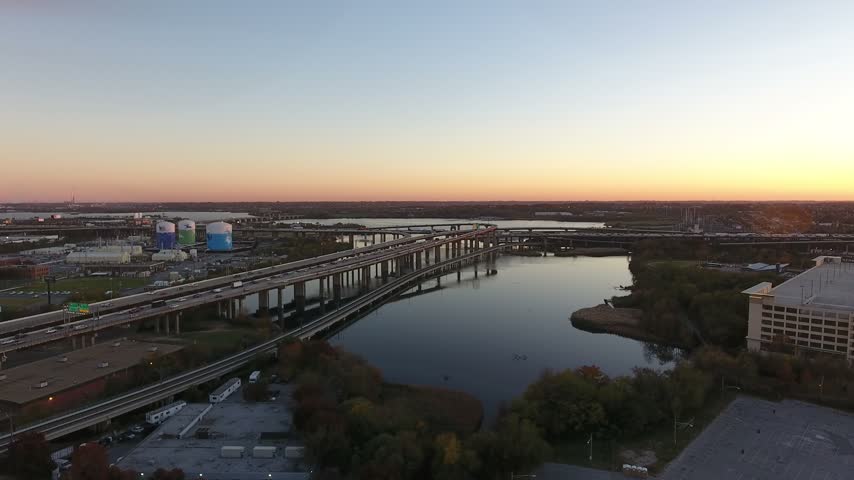 Aerial flight over the calm Middle Branch of the Patapsco River beside the elevated I-95 highway bridges at sunset, with traffic, autumn trees and an orange horizon. Baltimore, Maryland, USA.