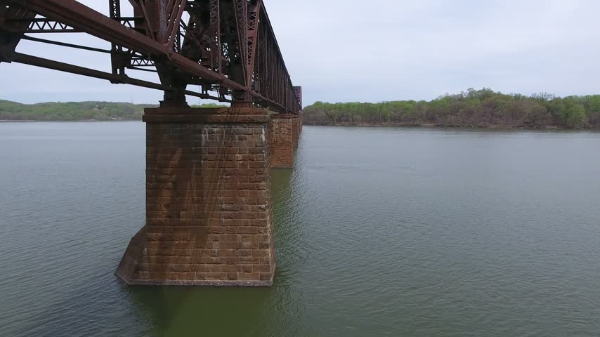 Aerial low glide past the stone piers beneath a steel railroad truss bridge over the calm Susquehanna River on an overcast spring day, with the far wooded shoreline. Havre de Grace, Maryland, USA.