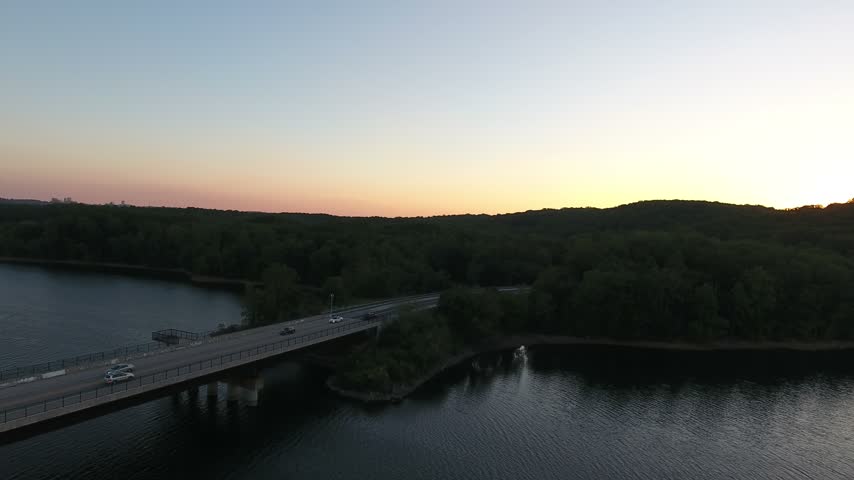 Aerial glide beside a highway bridge with traffic over Loch Raven Reservoir at dusk, with dark wooded hills and a pink and orange horizon reflected in calm water. Baltimore County, Maryland, USA.