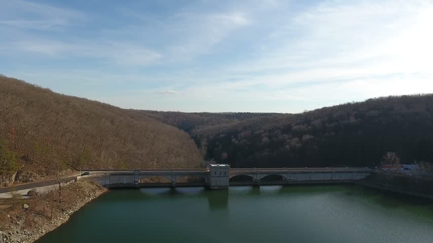 Aerial slow push across the green water of Prettyboy Reservoir toward the concrete Prettyboy Dam and gate tower between bare winter hills on a sunny day. Baltimore County, Maryland, USA.