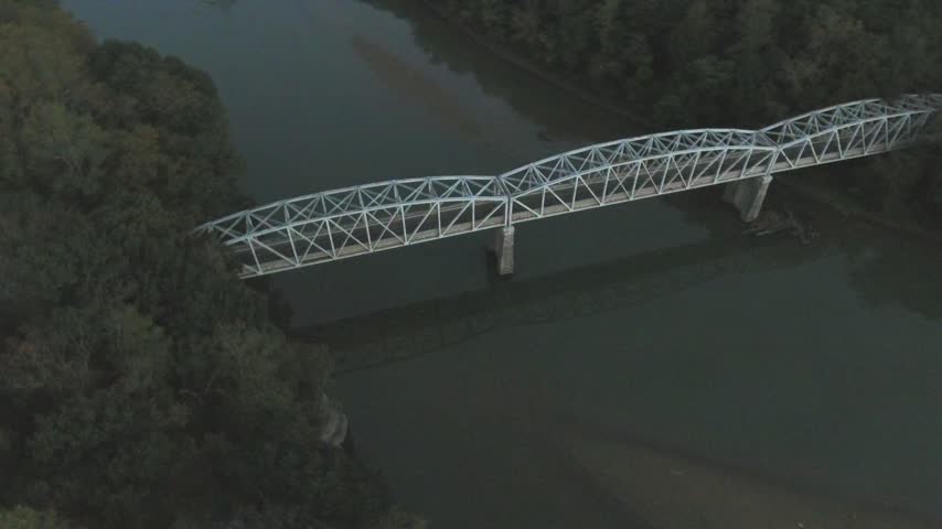 Aerial slow orbit around a white steel truss bridge on concrete piers spanning Loch Raven Reservoir at dusk, surrounded by dense forest and dark water. Cockeysville, Baltimore County, Maryland, USA.