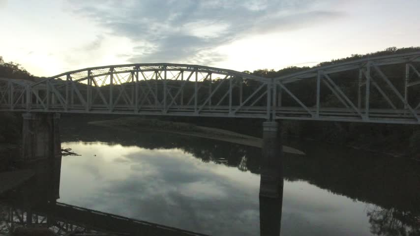 Aerial low pass near the water of Loch Raven Reservoir at dusk, with a steel truss bridge in silhouette and clouds reflected in the calm water between wooded hills. Cockeysville, Maryland, USA.