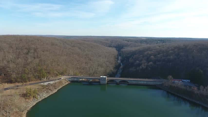 Aerial wide steady view over Prettyboy Reservoir with the long concrete Prettyboy Dam stretching across the valley between bare wooded winter hills under a blue sky. Baltimore County, Maryland, USA.