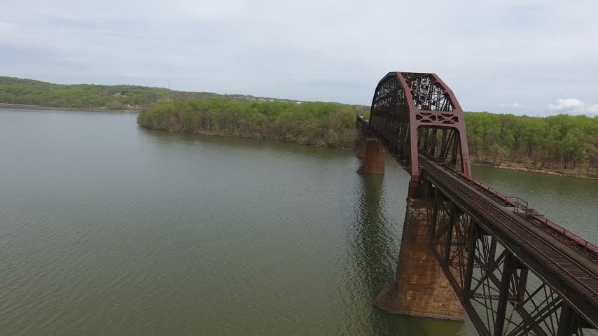 Aerial turn revealing the arched steel truss span of a railroad bridge on stone piers over the Susquehanna River on an overcast spring day, with green shorelines. Havre de Grace, Maryland, USA.