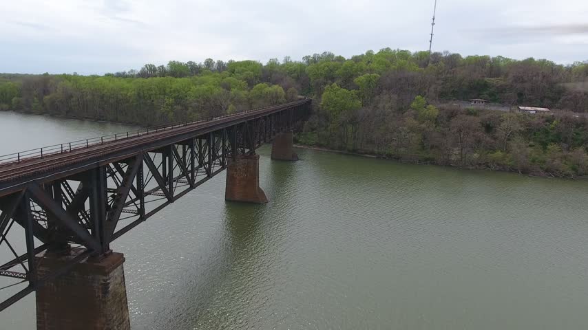Aerial sweep along a steel railroad truss bridge on stone piers over the Susquehanna River toward the far green wooded shore on an overcast spring day. Havre de Grace, Maryland, USA.