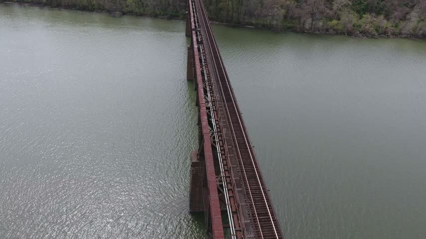 Aerial view of a steel railroad truss bridge deck running diagonally across the Susquehanna River toward a green wooded shoreline on an overcast spring day. Havre de Grace, Maryland, USA.