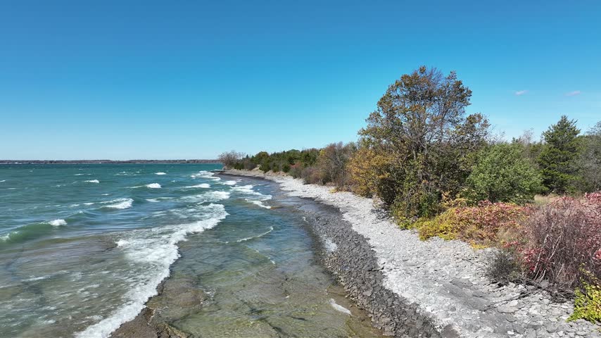 An aerial tracking shot glides along the rocky limestone shoreline of Lake Ontario as windblown turquoise waves crash into white foam beside autumn coastal trees under a clear blue sky in Prince Edward County, Ontario, Canada