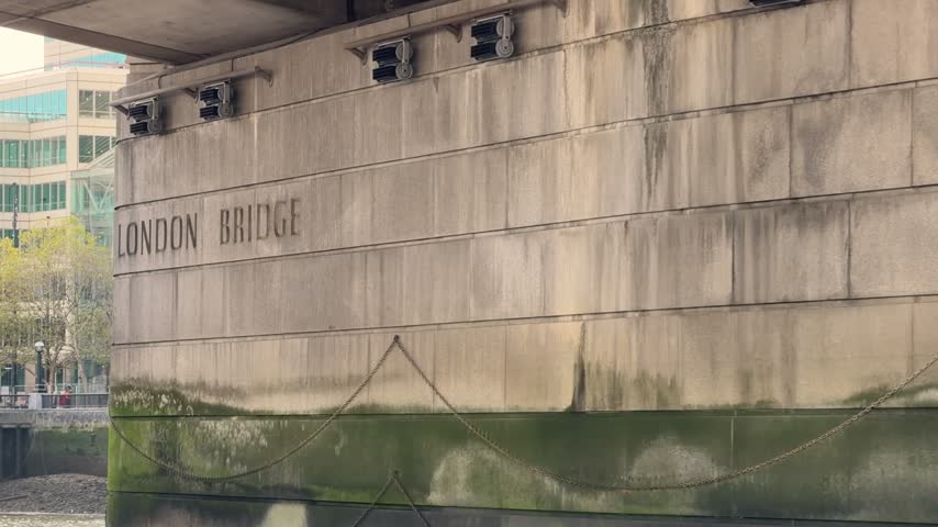 View from a passing boat traveling beneath London Bridge over the River Thames in London, England, showing the concrete bridge supports and historic landmark signage. 15518  