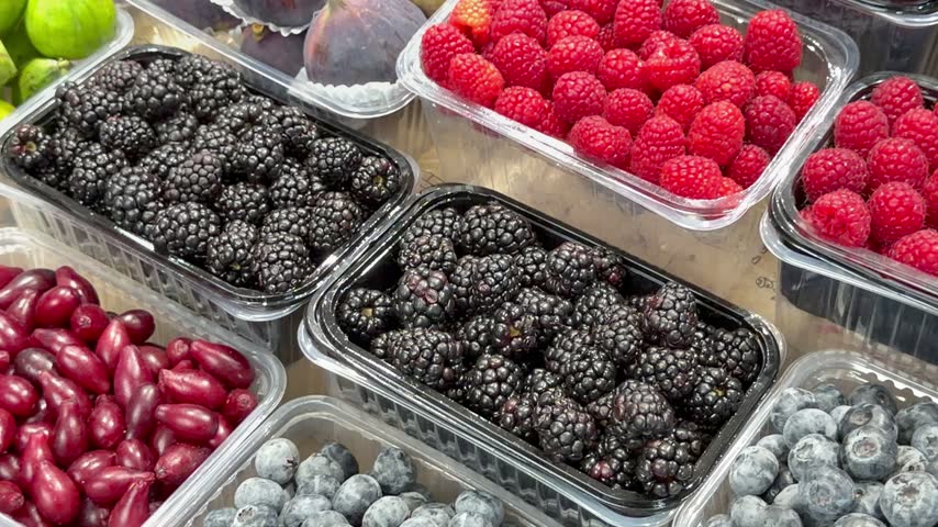 Fresh raspberries, blackberries, blueberries, and cornelian cherries packed in transparent plastic containers on market counter. Summer berry harvest, healthy food shopping, organic product display.