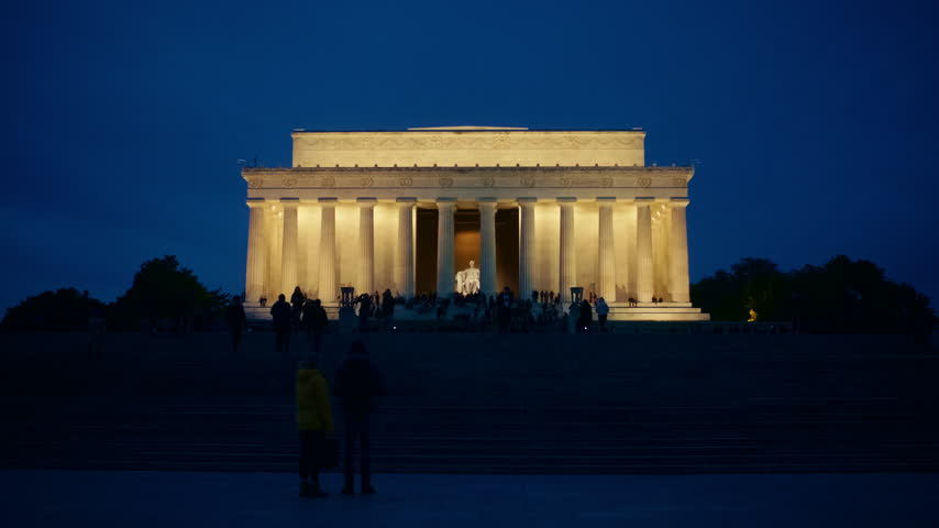 The illuminated Lincoln Memorial stands against a deep blue evening sky, with visitors on the steps and dark tree silhouettes beside the classical building. Filmed in Washington, DC, United States.
