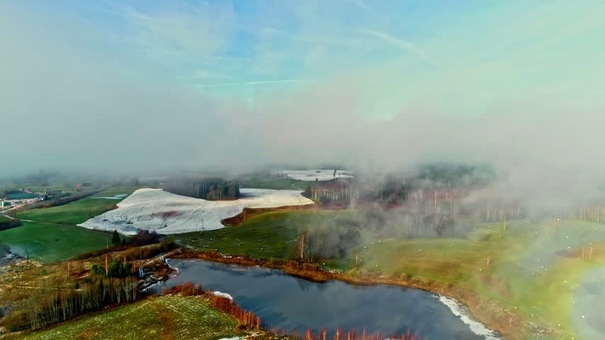 Aerial view of a small pond surrounded by green fields, patches of snow, and misty forests in Latvia.