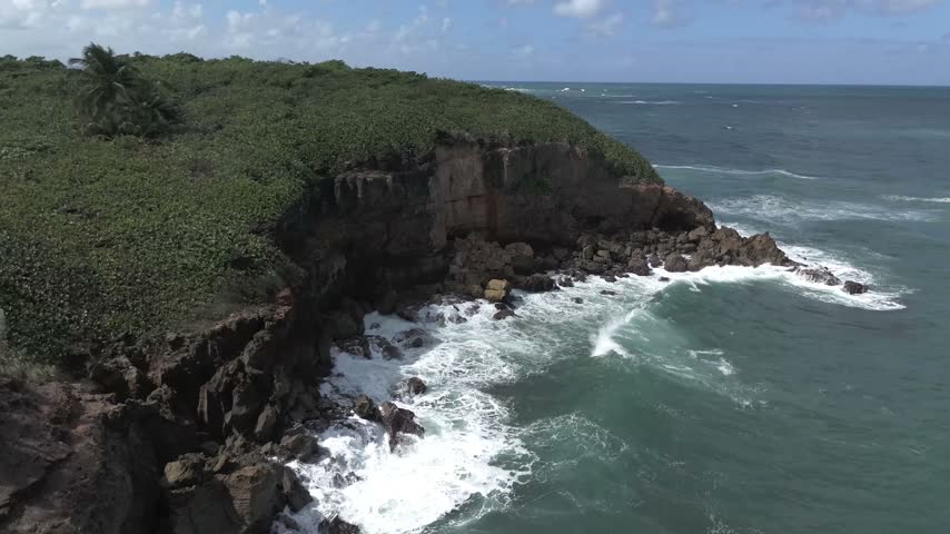 Low aerial view of red rock cliffs and green shrubs with white waves crashing on the rocky shore of Puerto Rico