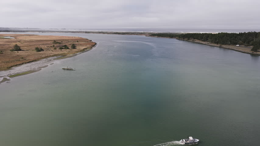 Salmon fishing boat travel the Coquille River in Bandon, Oregon, as anglers take part in the seasonal fishery across the broad coastal estuary. Drone pullback shot with ocean in the distance.