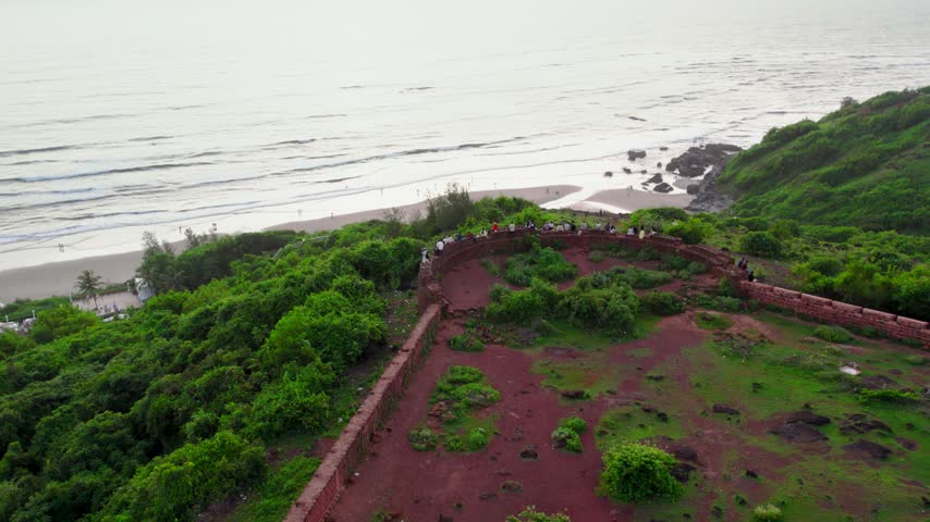 Aerial view of historic fort walls overlooking the sandy coastline and the Arabian Sea with people gathered along the ramparts in Fatrade, Goa, India.