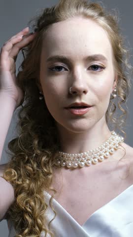 Blonde woman in bridal dress poses gracefully while adjusting pearl necklace. Charming female model reveals calm expression and flawless skin during studio photography. Рair is gathered into a braid.