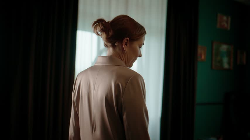 Woman in a beige blazer stands near a window, gazing out thoughtfully. Her hair is pulled back into a neat bun. Soft natural light filters through sheer curtains. The room features dark curtains