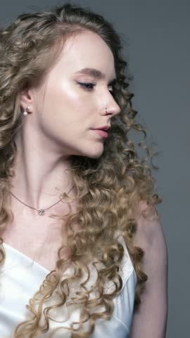 Young woman with long curly hair turns gently during bridal beauty session. Calm female model displays elegant wedding hairstyle while standing against plain background.