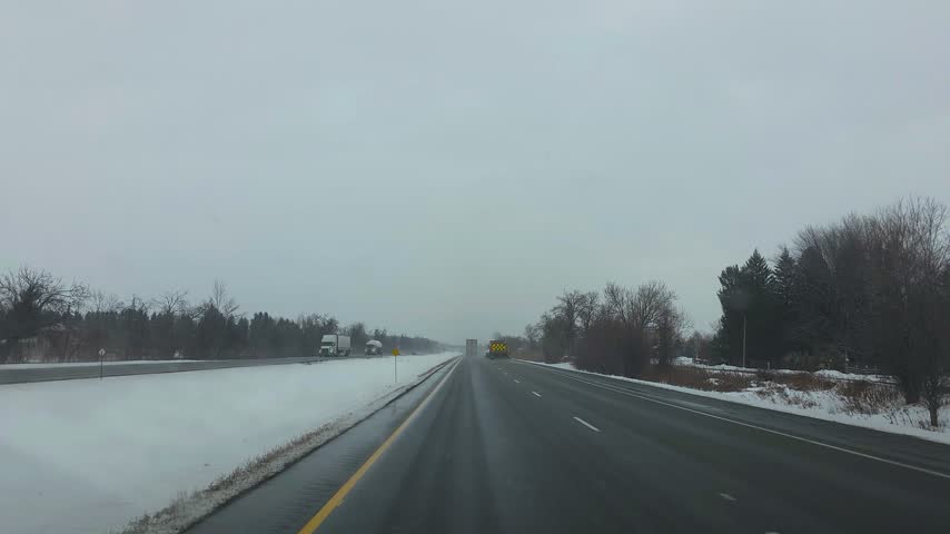 A vehicle travels along a snowy highway past a maintenance truck and bare trees.
