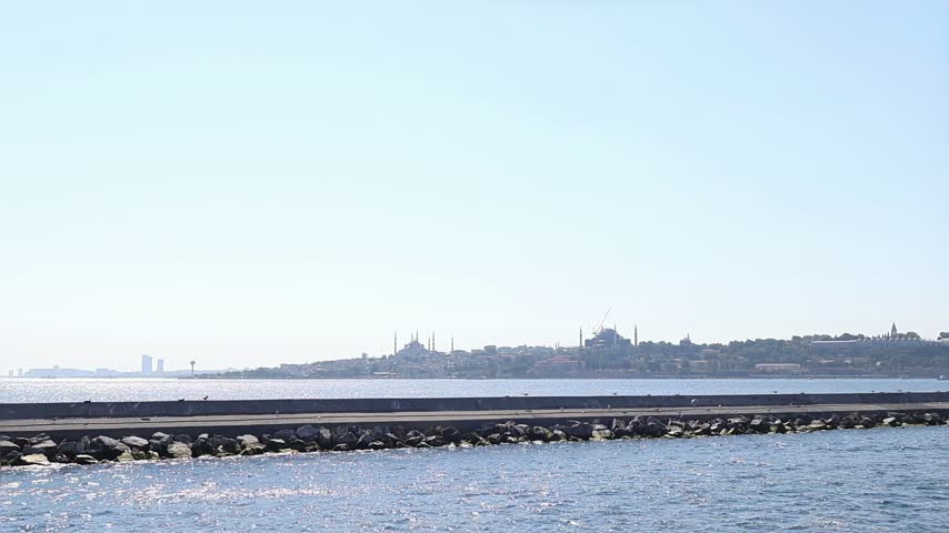 Panoramic view of Istanbul, Turkey, across blue water with a rocky breakwater in foreground and historic city skyline with minarets visible in distance. Peaceful travel and coastal city background