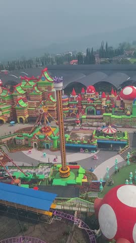 Aerial view of whimsical theme park with colorful structures. Large mushroom and castle-like buildings dominate the scene. Tall tower ride stands near a twisting roller coaster track