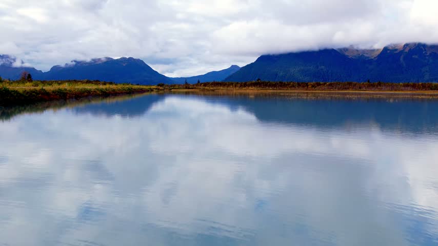 Low drone flight skimming a glassy wetland pond with perfect mirror reflections of clouds and low cloud covered Chugach Mountains near Cordova Alaska. 4K 60fps aerial footage.