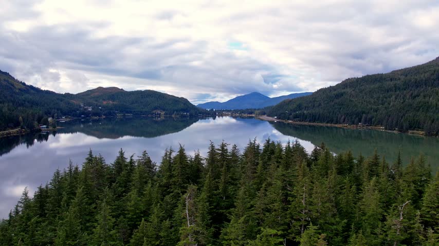 Drone rises over spruce treetops to reveal glassy Eyak Lake mirroring clouds between forested hills and distant mountains near Cordova Alaska. 4K 60fps aerial reveal footage.