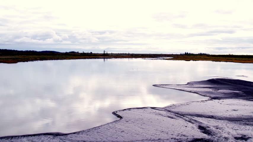 Low drone flyover past silty mudflats and across bright reflective wetland water toward distant swans on the flat marsh horizon near Cordova Alaska. 4K 60fps aerial footage.