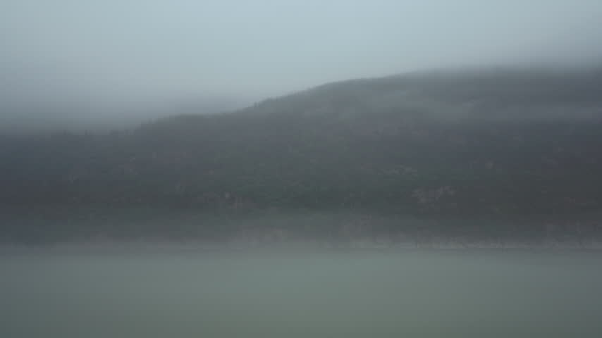 Fog drifting over a forested shore with a distant waterfall