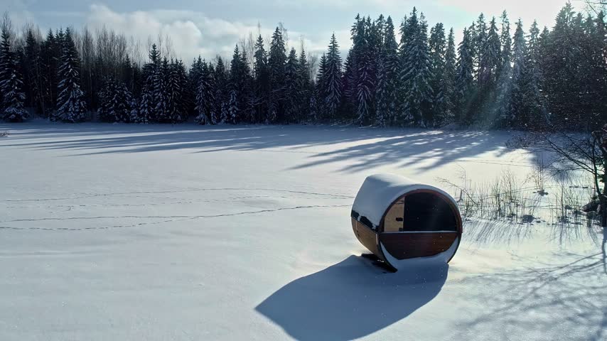 Aerial view of a barrel sauna sitting in a snow-covered field near a dense forest in Latvia.