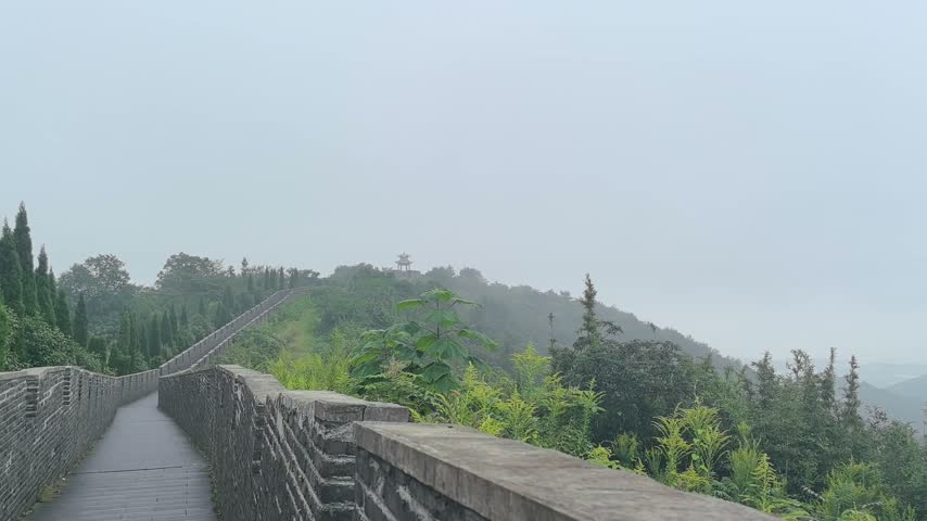 The camera moves forward along a long gray brick walkway extending across a lush green hillside in misty weather.