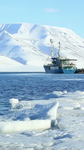 A ship anchored in the blue waters of the Arctic Ocean near sea ice with snow-covered mountains in the background in Svalbard Norway