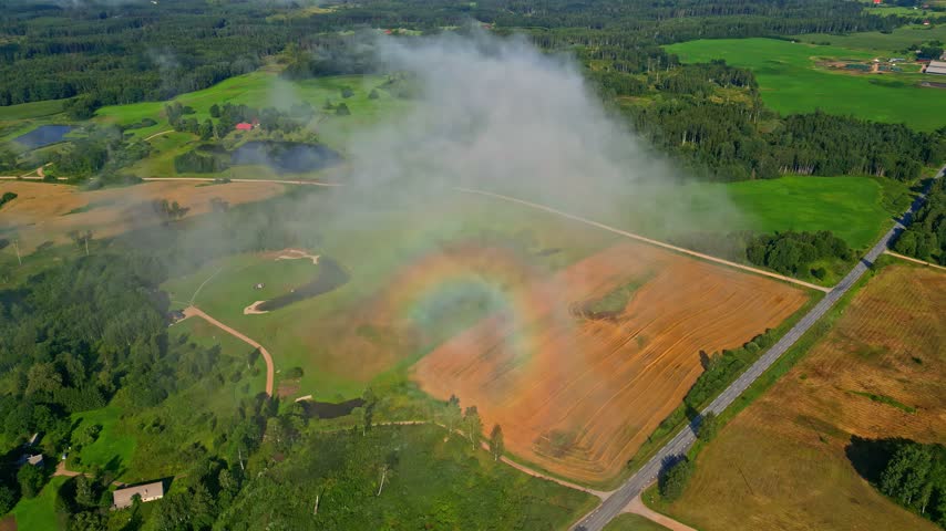Aerial view of a vibrant rainbow appearing through low-hanging fog over green and golden agricultural fields in Latvia.