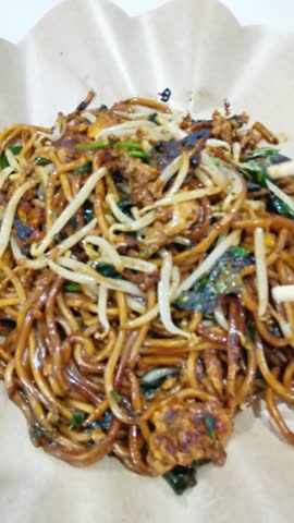 A close-up shot of a savory Asian noodle dish, likely fried noodles with bean sprouts and vegetables, served in a paper bowl and being lifted with chopsticks.