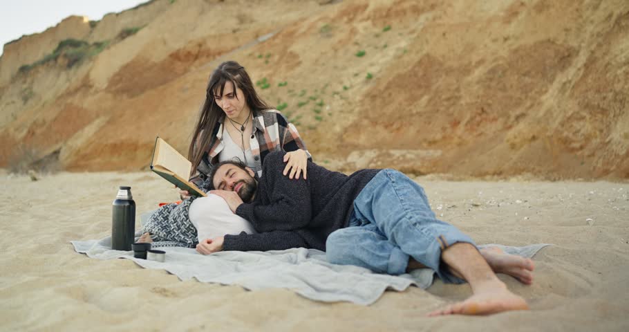 People relaxing on sandy beach near cliff rocks, woman reading book and adjusting sweater and hair while man reclines with head in her lap in warm daylight and calm