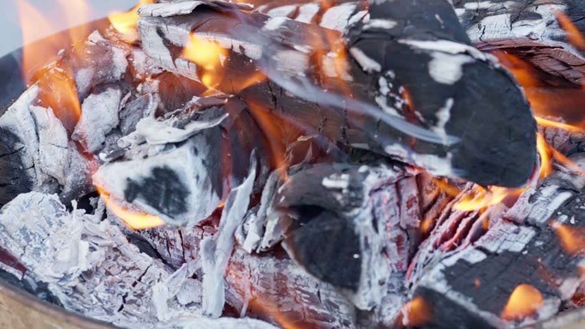 Close-up of wooden logs burning with bright flames and turning into hot glowing embers in an outdoor fire pit or grill, creating a warm atmosphere