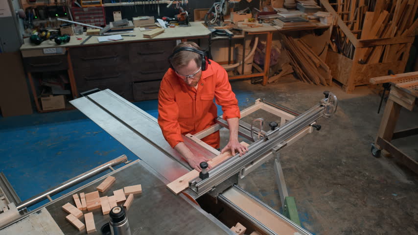 Caucasian middle aged carpenter cutting wooden board using sliding table saw in prison workshop. Footage suitable for industrial woodworking tutorials, carpentry training, manufacturing marketing
