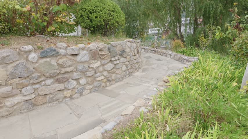Stone walkway curves beside rustic retaining wall through lush garden landscape, peaceful outdoor pathway with willow greenery, lake views, ornamental grass, and tranquil China travel scenery
