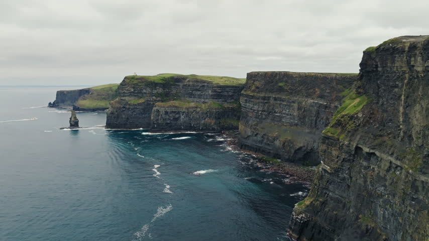 Overcast aerial view of layered sea cliffs at Cliffs of Moher in County Clare, Ireland. A rock stack and white wave foam mark this Atlantic coast sightseeing landscape.