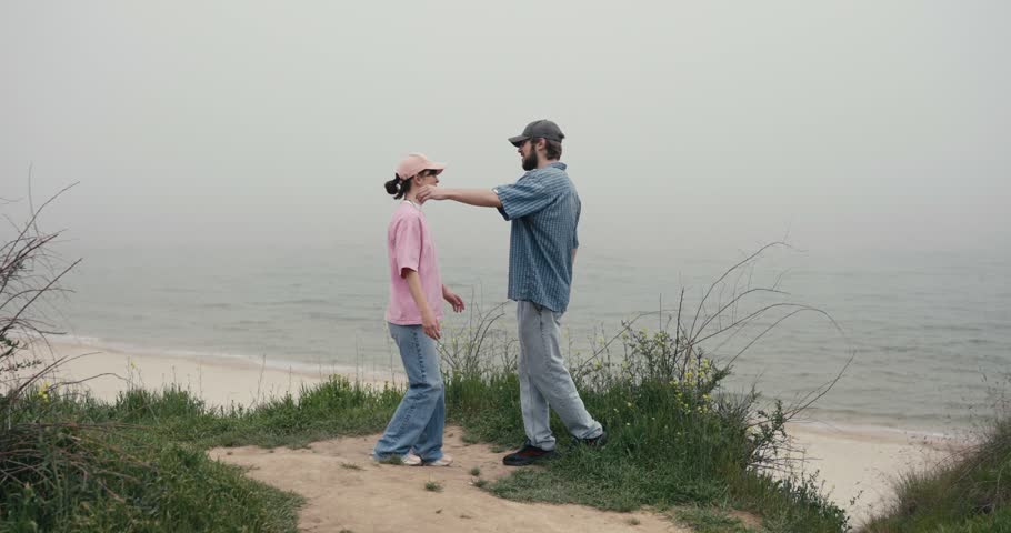 Two people embracing on foggy coastal bluff above calm ocean, pink shirt cap and blue checked shirt, playful leg lift on grassy overlook Esrda afdsadfadsafsdafadsfadsf
