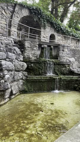 Cascading waterfall fountain in stone grotto at park with bridge.
Peaceful landscape featuring an artificial multi-tiered water cascade built from natural rock. Includes a small pond and hanging willow branches. Perfect for landscaping concepts, park designs, and zen relaxation videos.