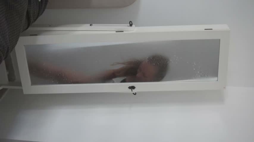 Young woman wipes mirrored cabinet with cloth in clean white bathroom, creating smooth reflective streaks during routine household cleaning, soft indoor light, modern minimalist interior.