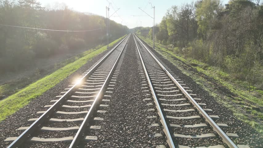 Train tracks stretch into the distance through a lush green landscape, showcasing the gradual transition of scenery along the railway corridor under a clear blue sky